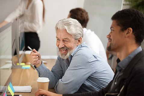 En äldre medarbetare sitter med en yngre medarbetare och samtalar vid en arbetsplats.